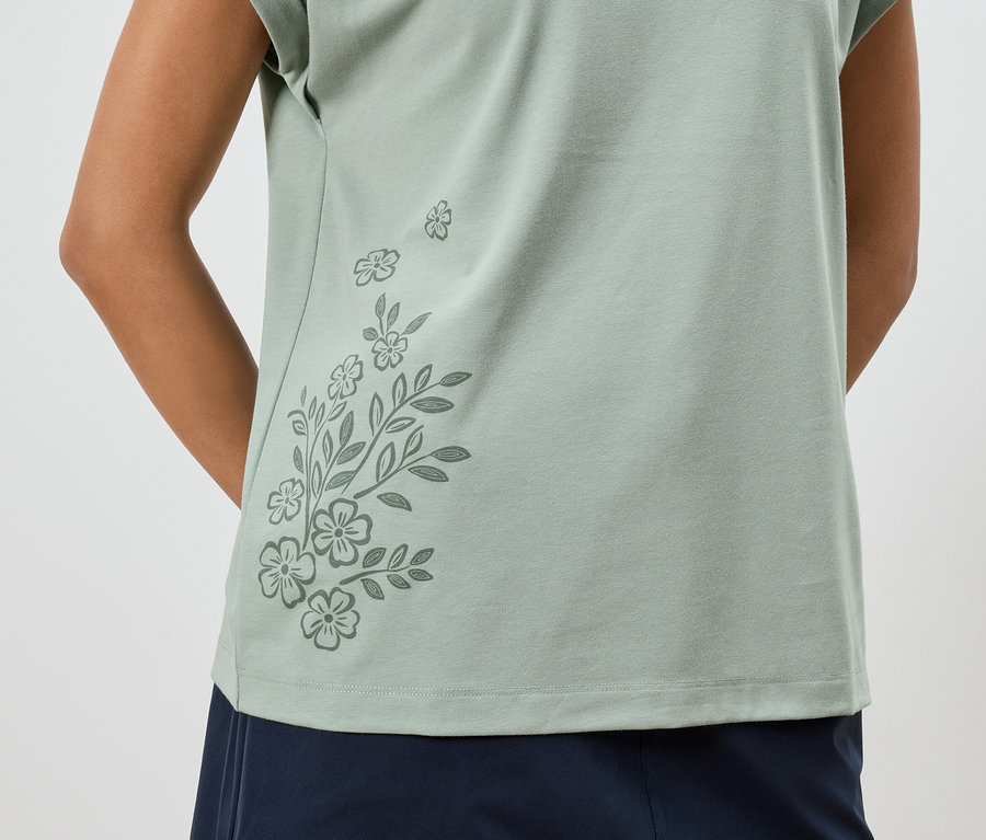 Nahaufnahme einer Frau in einem hellgrünen T-Shirt mit Blumenmuster und einer dunkelblauen Hose.