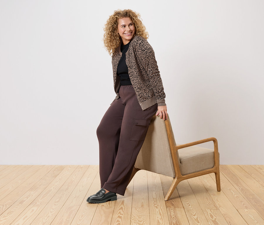 Frau mit lockigem Haar lehnt an einem Sessel, trägt eine braune Hose, einen schwarzen Pullover und eine braun-schwarze Strickjacke mit Tiermuster.