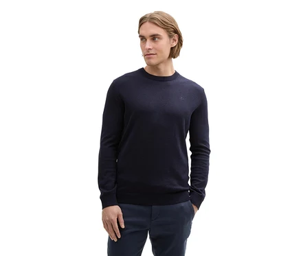 Mann mit blondem Haar, der einen dunkelblauen Rundhals-Pullover mit kleinem Logo auf der Brust und eine dunkelblaue Hose trägt. Seine linke Hand steckt in der Hosentasche, und er blickt nach rechts.