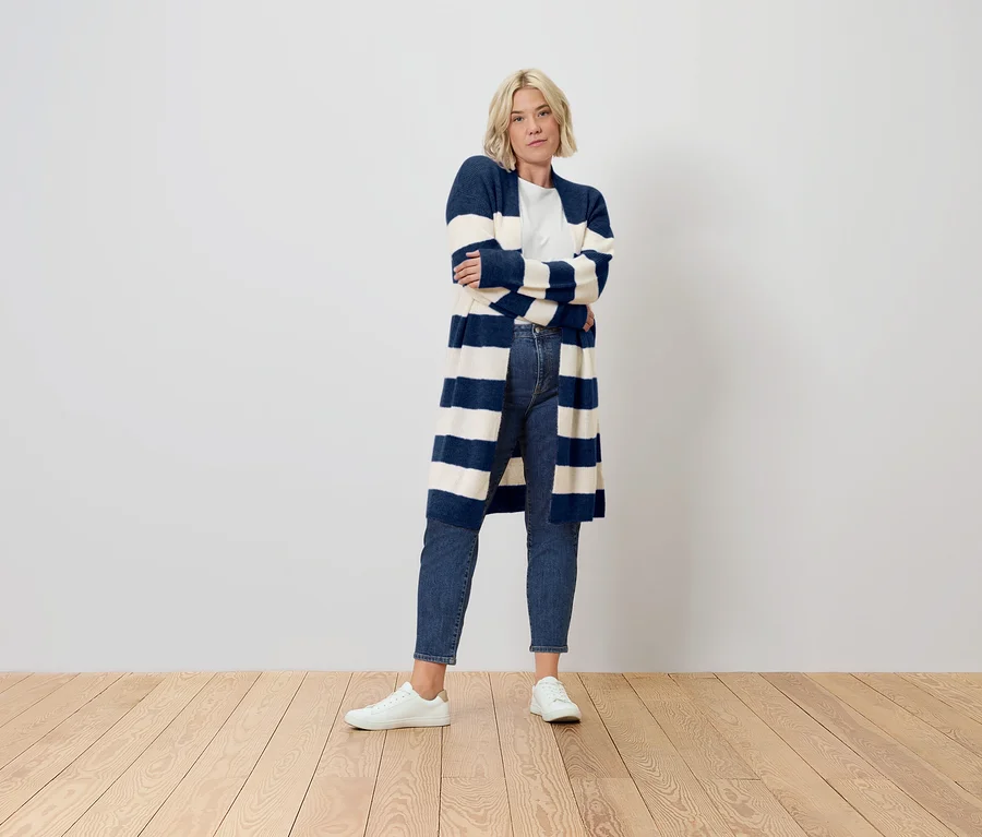 Blonde Frau mit verschränkten Armen, die eine lange, blau-weiß gestreifte Strickjacke, Jeans und weiße Sneaker trägt.