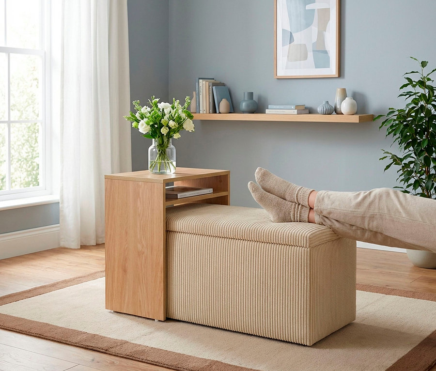 Beine einer Person mit hellen Socken ruhen auf einem beigen, gerippten Ottomanen. Daneben steht ein Holztisch mit einer Vase weißer Blumen. Der Raum hat eine blaue Wand mit einem Regal, ein Fenster mit Vorhängen und eine Topfpflanze.