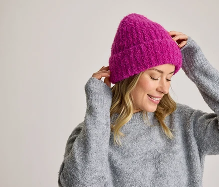 Lächelnde Frau in einem grauen Pullover, die eine pinkfarbene Strickmütze zurechtrückt.
