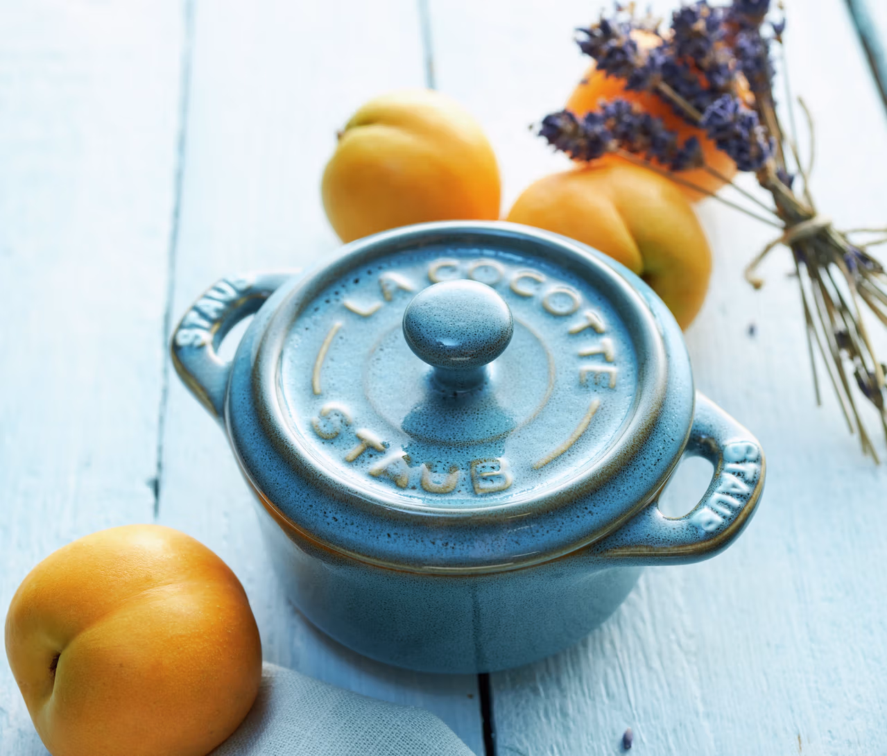 Antiktürkise STAUB Keramik-Cocotte mit Deckel, umgeben von Aprikosen und einem Lavendelstrauß auf hellem Holz.