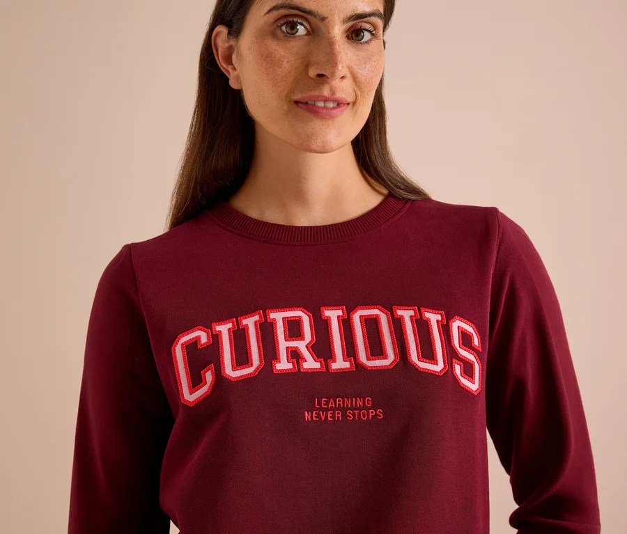 Frau trägt ein bordeauxrotes CECIL Sweatshirt mit der Aufschrift „CURIOUS“ und „LEARNING NEVER STOPS“.
