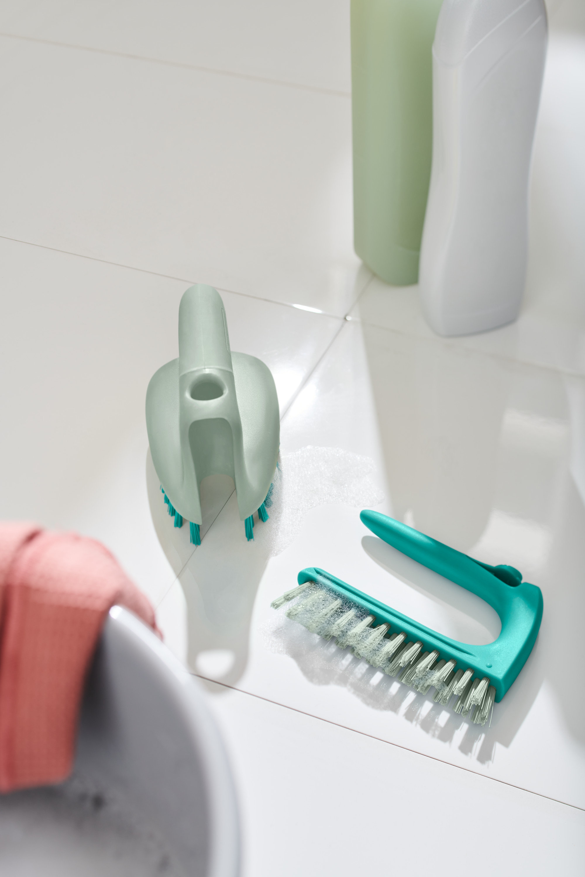 Zwei 2-in-1-Fliesen-Bodenbürsten liegen auf weißen Fliesen neben einem Waschbecken.