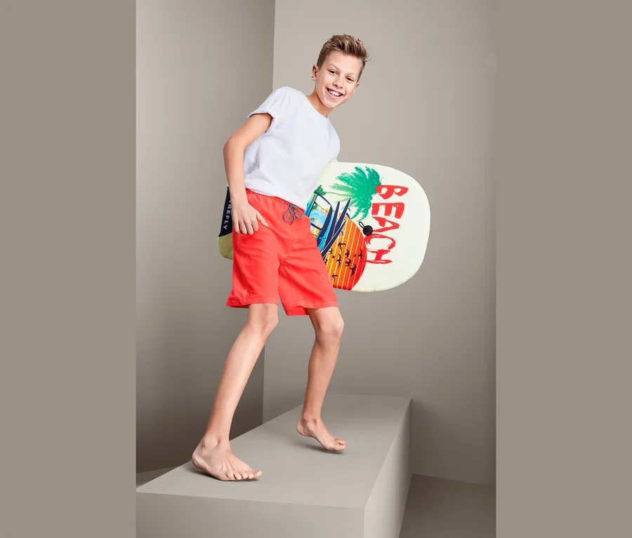 Ein Junge in roten Boardshorts hält ein Brett mit Strandmotiv.