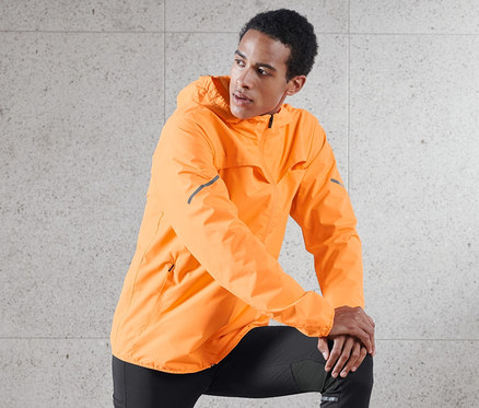 Mann posiert mit einem Bein auf einem Holzhocker, trägt eine neonorange Windprotection-Jacke.