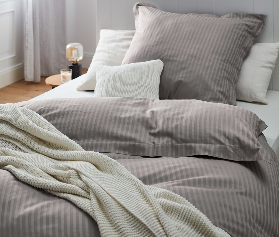 Nahaufnahme eines Bettes mit grauer Premium-Satin-Bettwäsche in verschiedenen Größen und einer weißen Strickdecke.