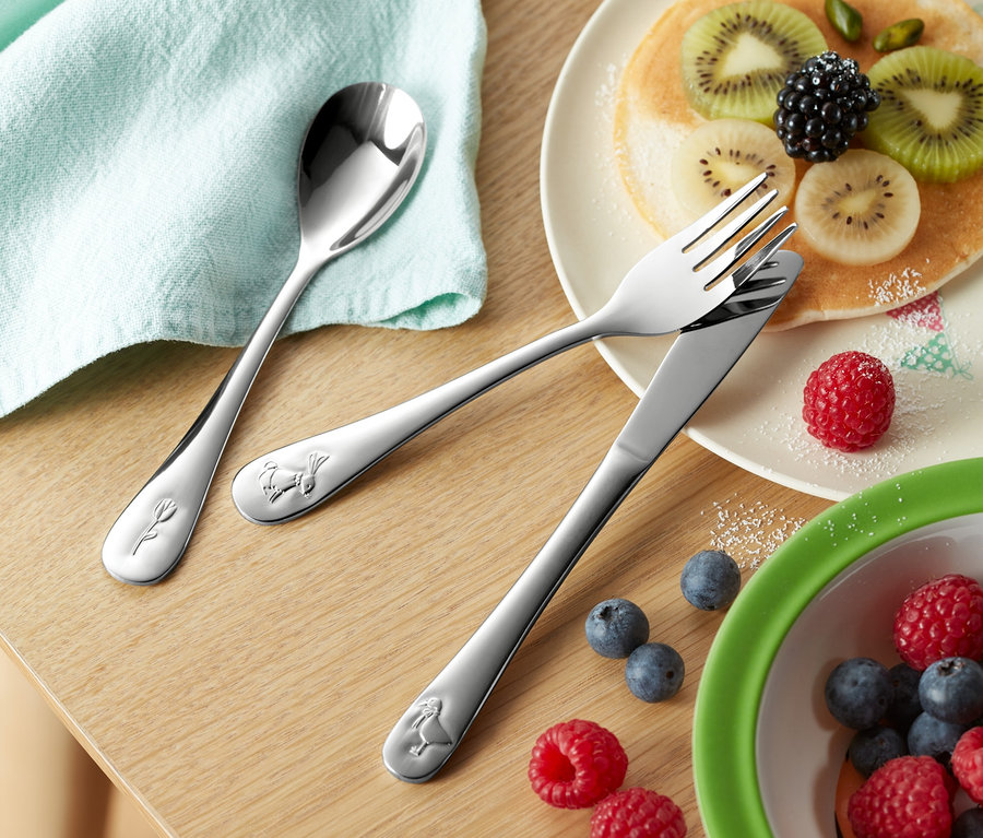 Auf einem Holztisch liegen ein Kinder-Besteck-Set (3-teilig), ein Teller mit Pfannkuchen und Früchten, eine Schüssel mit Blaubeeren und Himbeeren und ein Glas mit Getränk.