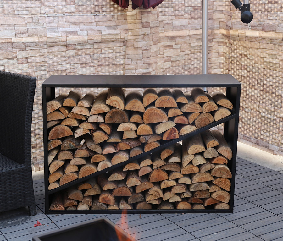 Ein ca. 114 cm großes Kaminholzregal aus schwarzem Metall ist mit Holzscheiten gefüllt. Im Hintergrund befindet sich ein Korbsessel.