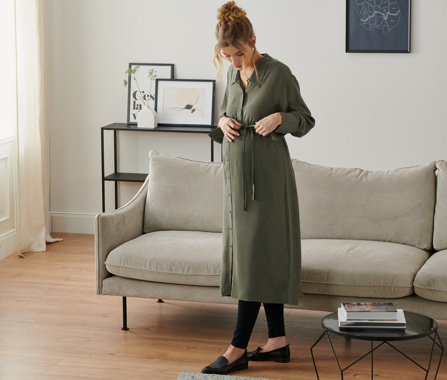 Eine schwangere Frau bindet ein olivgrünes Umstands-Blusenkleid in einem Wohnzimmer.