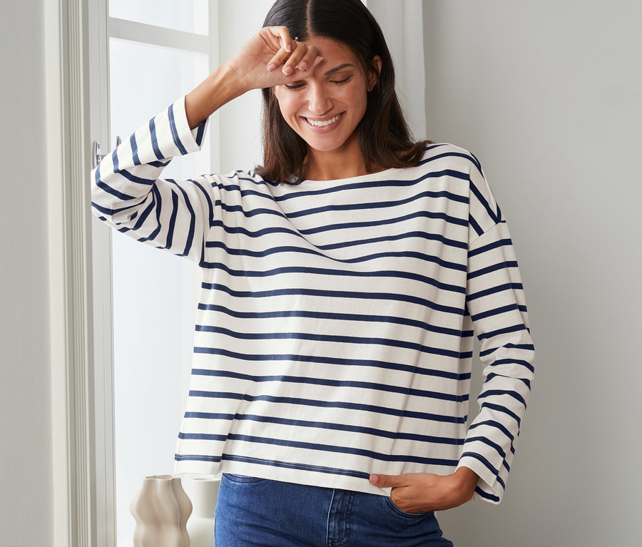 Eine Frau lächelt und steht am Fenster und trägt ein gestreiftes Sweatshirt.