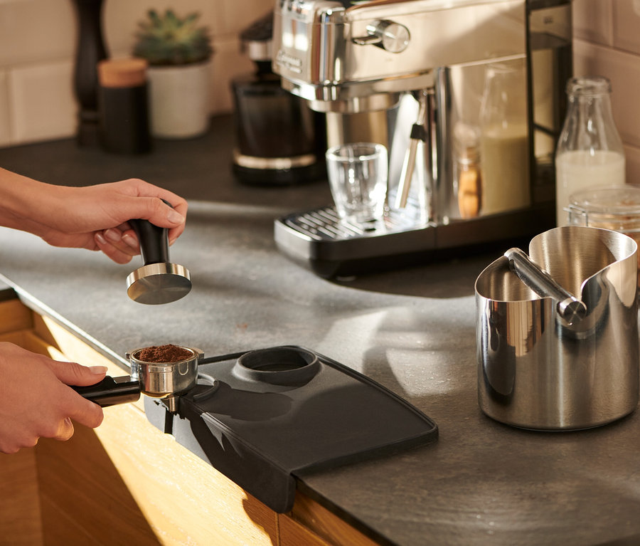 Eine Frau tampert Kaffee auf einer schwarzen Tampermatte.