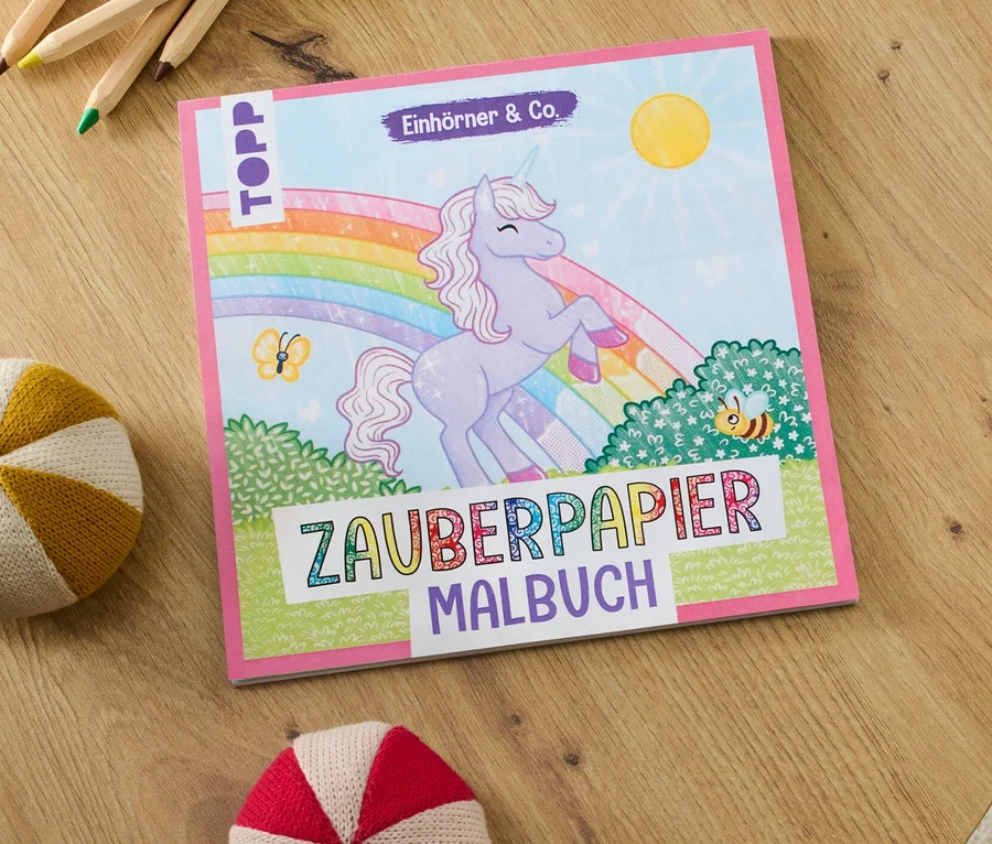Auf einem Tisch liegen das Buch "Zauberpapier Malbuch ‒ Einhörner & Co.", Buntstifte und zwei Strickbälle.