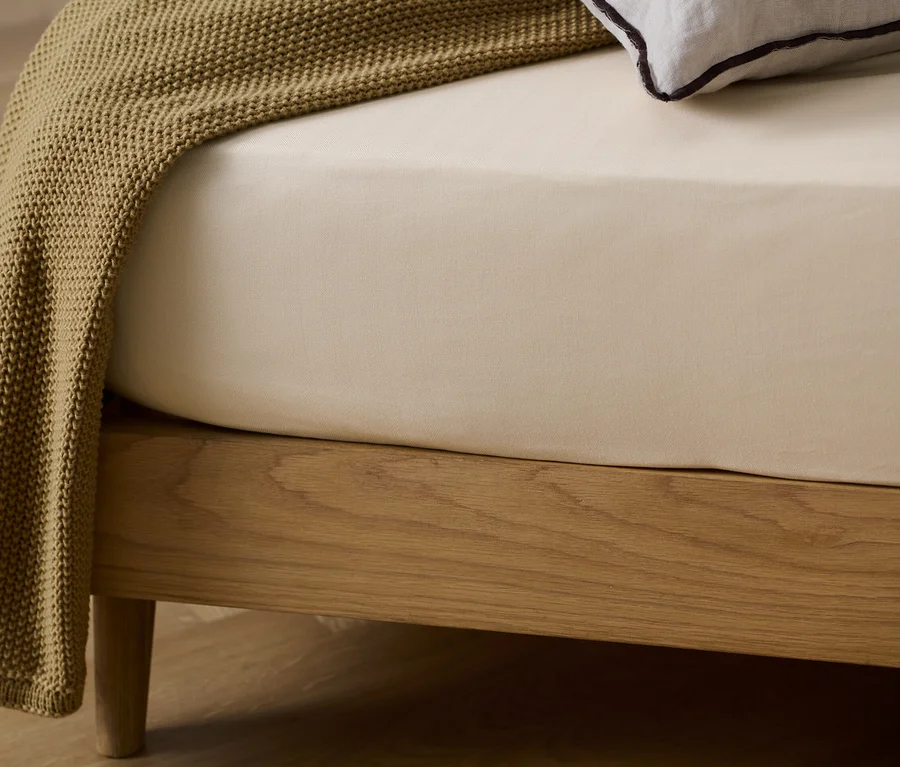 Detailansicht eines Holzbettes mit einem beigen Edelflanell-Spannbettlaken und einer braunen Strickdecke.