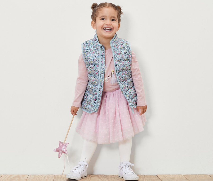 Lächelndes Mädchen trägt eine geblümte Kinder-Steppweste, ein rosa Langarmshirt, einen rosa Rock und weiße Turnschuhe und hält einen Zauberstab.