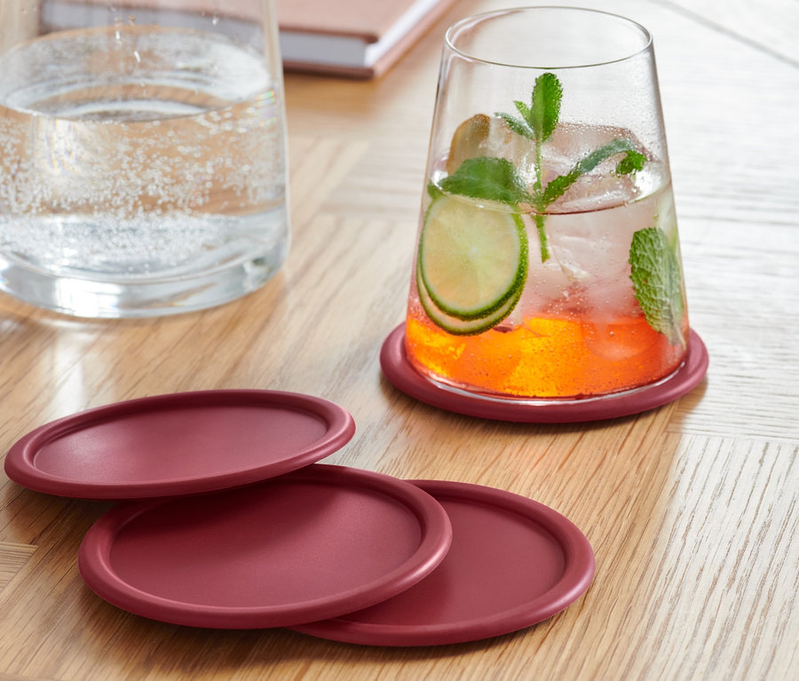 Ein mit einem Getränk gefülltes Glas steht auf einem der vier bordeauxroten Glasuntersetzer. Drei weitere Untersetzer liegen auf einem Holztisch. Im Hintergrund stehen eine Wasserkaraffe und ein Notizbuch.