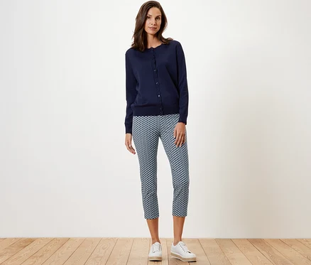 Frau steht und trägt eine bedruckte 7/8-Stretchhose, eine marineblaue Strickjacke und weisse Turnschuhe.