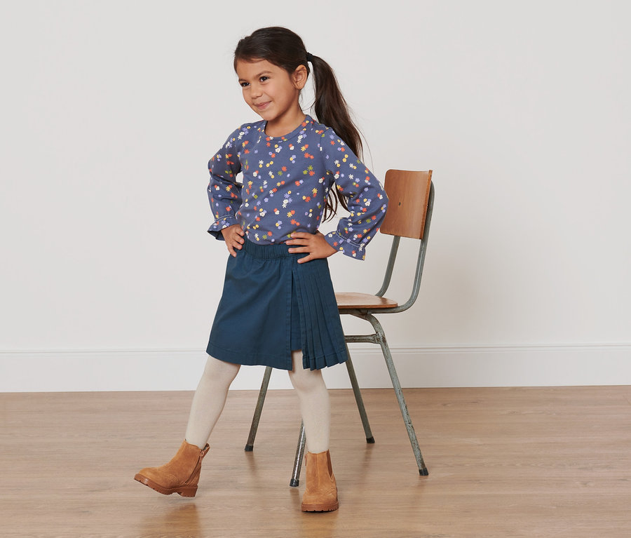 Mädchen posiert in blauer Bluse mit Blumenmuster und dunkelblauem Faltenrock. Sie trägt einen Kleinkinder-Kiltrock.