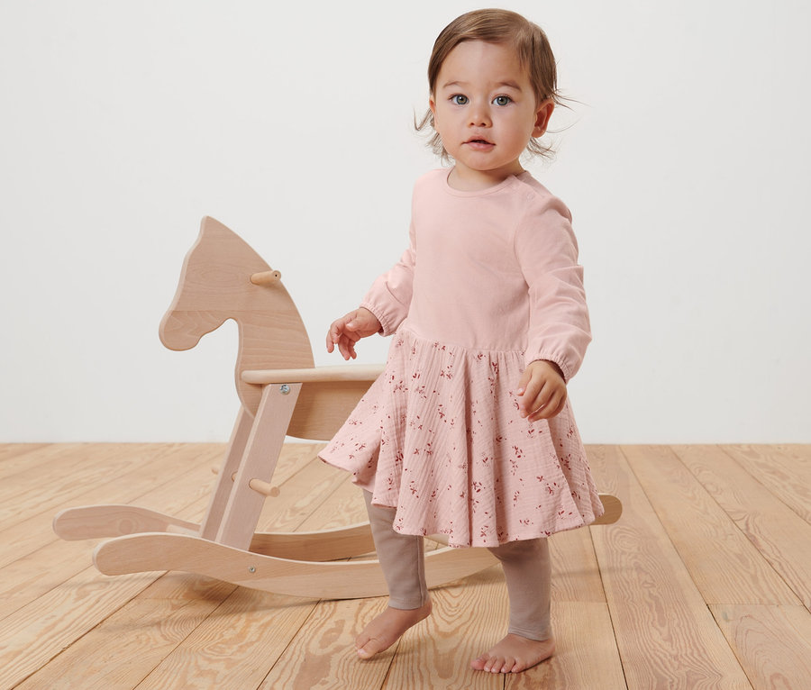 Kleinkind steht neben einem Holzschaukelpferd und trägt ein rosa Webkleid.