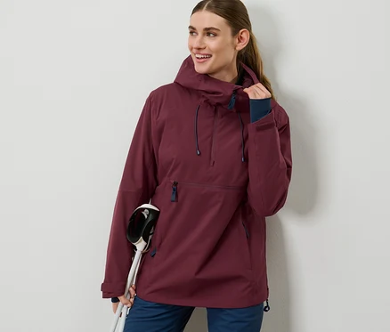 Frau trägt eine bordeauxrote Halfzip-Skijacke und hält Skistöcke.