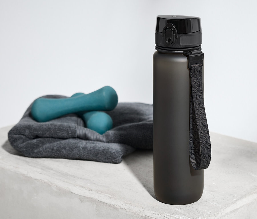 Eine Trinkflasche steht auf einer grauen Oberfläche neben einem Handtuch und Hanteln.
