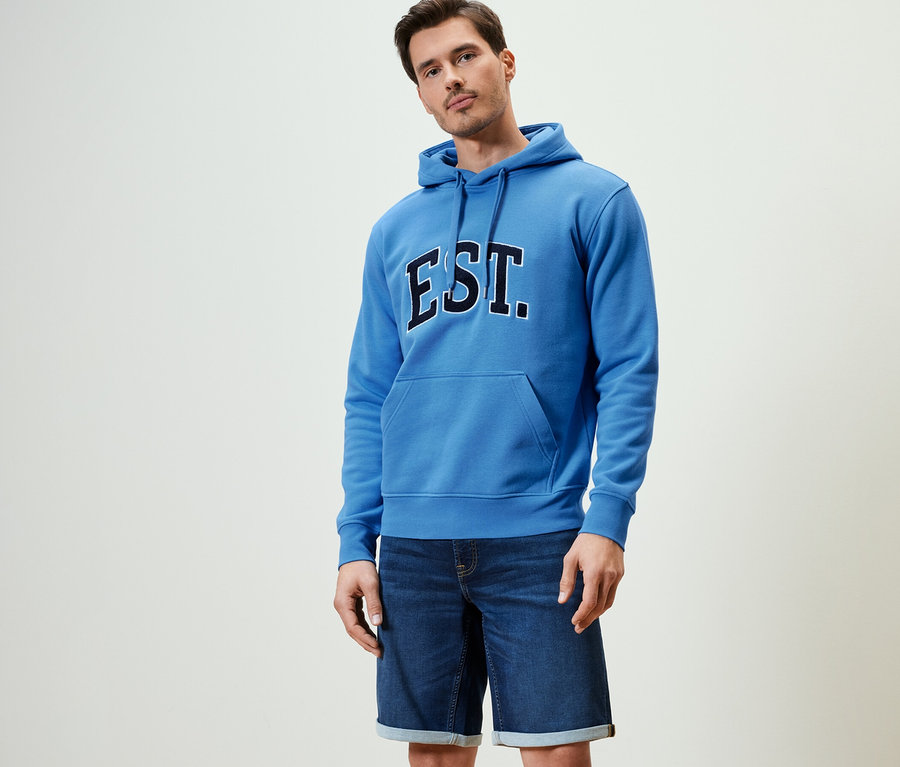 Mann posiert in blauem Hoodie und Jeansshorts.