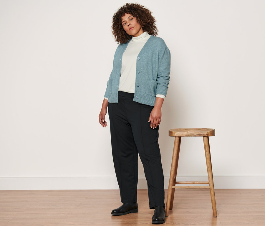 Frau, die neben einem Holzhocker steht, trägt eine Strickjacke mit Wolle, ein cremefarbenes Rollkragenshirt, eine schwarze Jerseyhose mit Biese und Lederboots.