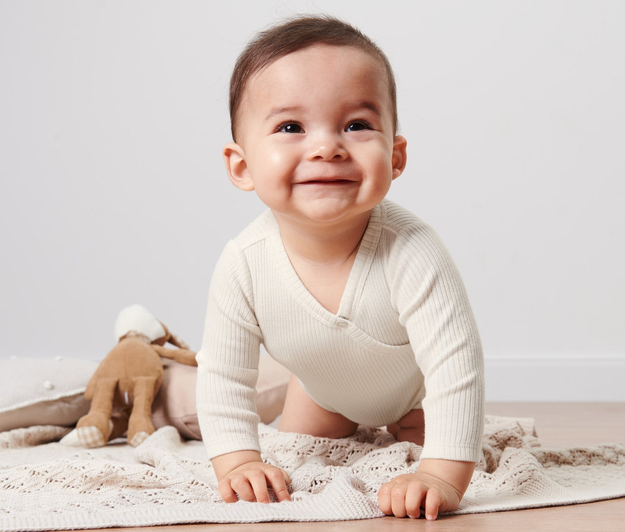 Lächelndes Baby im Baby-Body mit Modal krabbelt auf einer beige Strickdecke neben einem Hasen-Spielzeug.