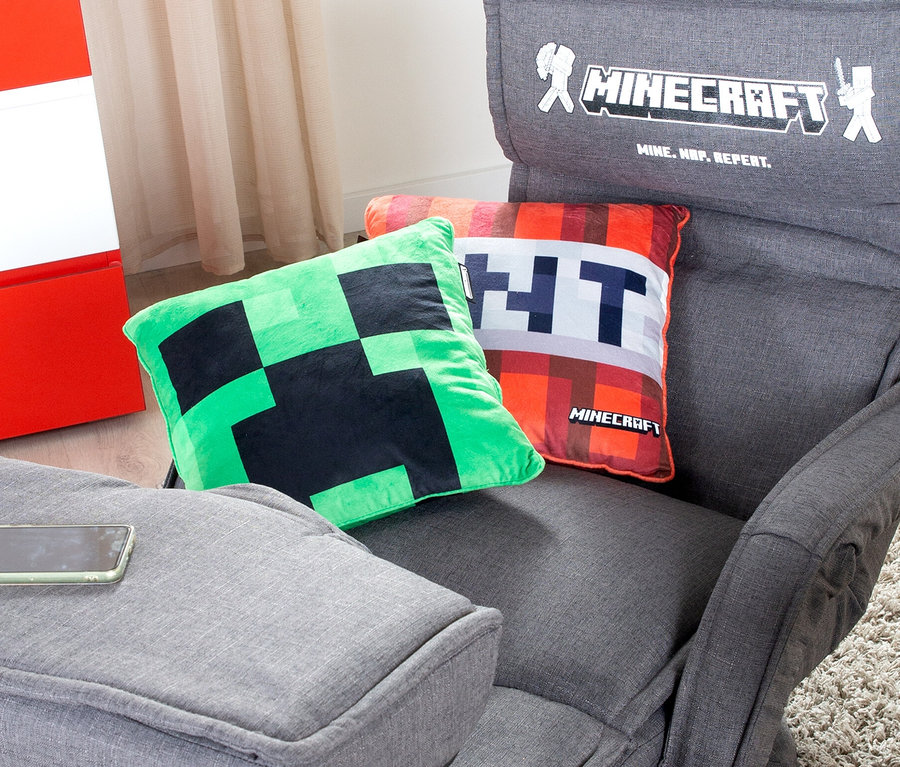Auf einem grauen Sessel liegen zwei Kissen, eines grün mit schwarzem Muster und eines orange mit der Aufschrift TNT. Auf der Rückenlehne des Sessels befindet sich das Minecraft-Logo.