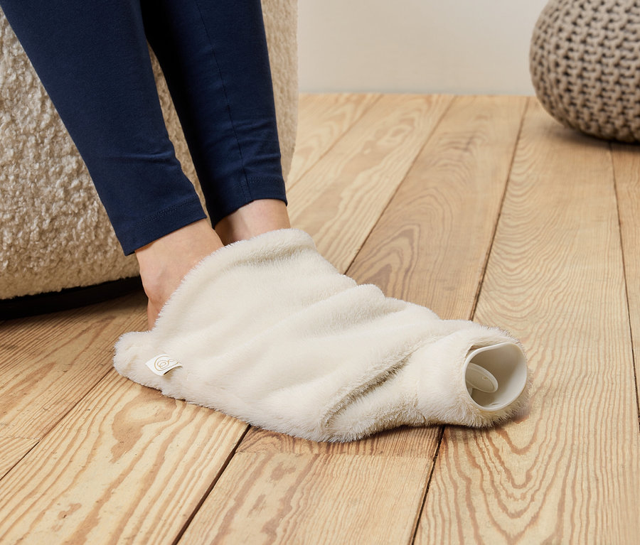 Füße in blauen Leggings stecken in einem flauschigen, cremefarbenen Wärmflaschenüberzug.