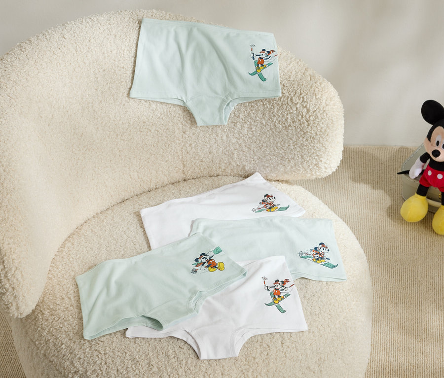 Auf einem flauschigen Sessel liegen vier Kinderunterhosen mit einem Bild von Mickey Mouse auf Skiern, und neben dem Sessel sitzt ein Plüsch-Mickey Mouse.