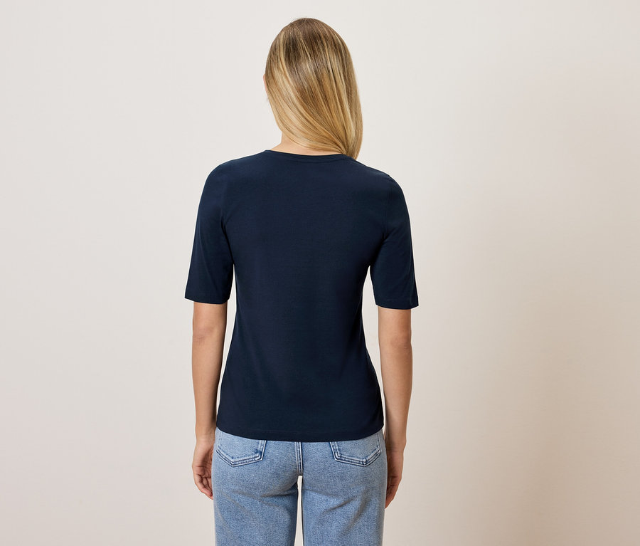 Eine Frau mit blonden Haaren, von hinten gesehen, trägt ein dunkelblaues T-Shirt und blaue Jeans.