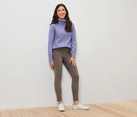 Frau steht und trägt einen lila Rollkragenpullover, braune Leggings und weiße Turnschuhe.