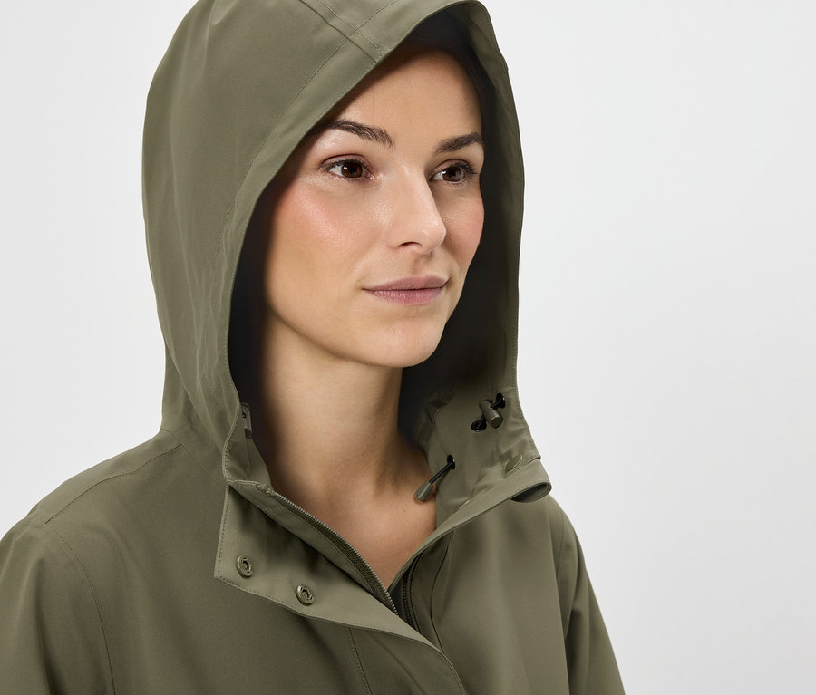 Frau mit braunen Haaren trägt eine olivgrüne Jacke mit Kapuze.