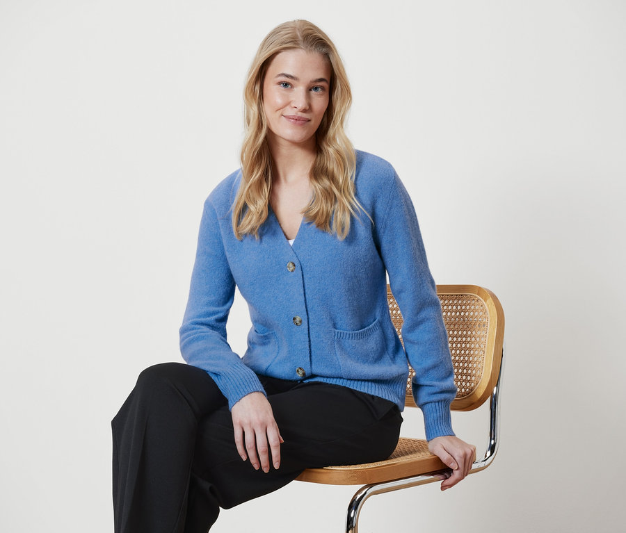 Blonde Frau sitzt auf einem Stuhl und trägt einen blauen Cardigan und eine schwarze Hose.