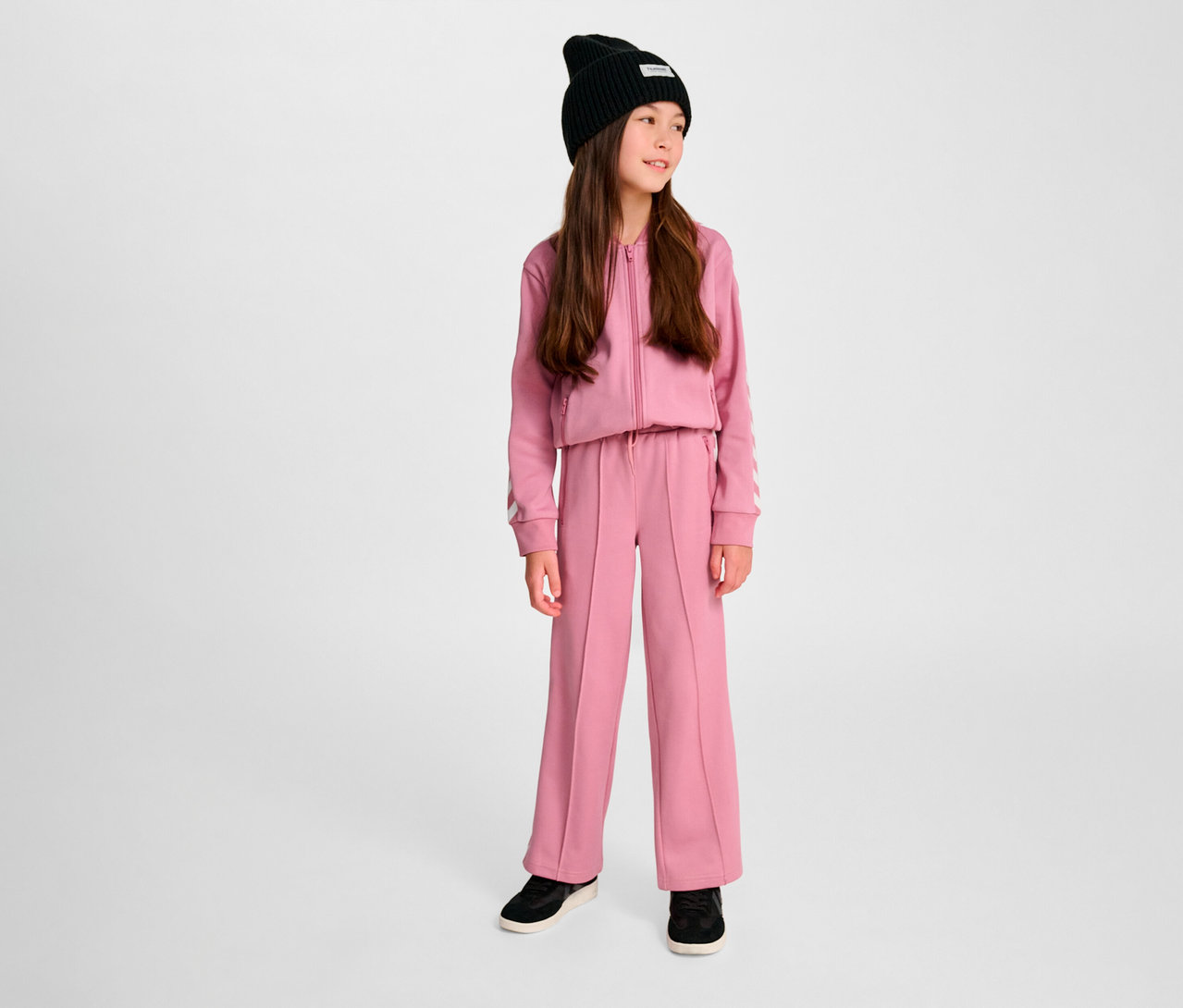 Mädchen trägt Trainingsanzug in Pink und schwarzer Mütze. Sie trägt HUMMEL HMLJR Archive Wideleg Poly Pants.