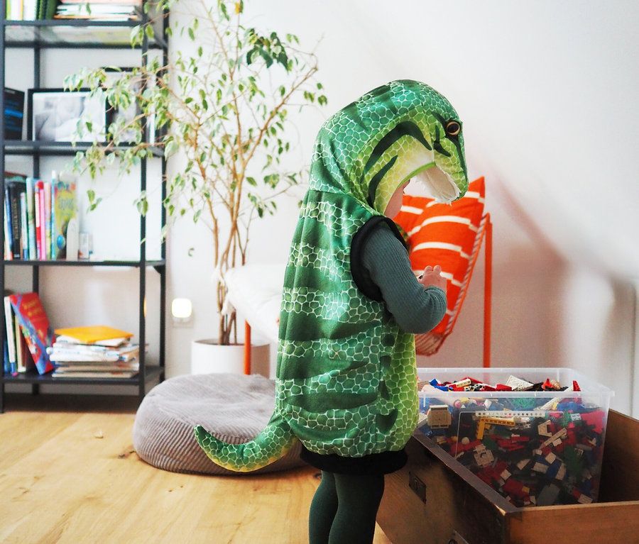 Kind im Dinosaurier-Kostüm steht neben einer Spielzeugkiste und hält ein orangefarbenes Kissen. Es trägt eine small foot Kostüm-Weste »Dinosaurier«.