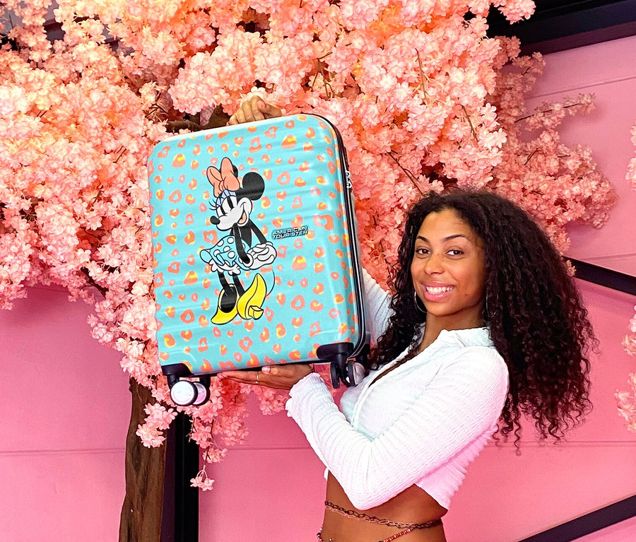Junge Frau hält einen kleinen American Tourister Wavebreaker »Minnie Mouse« Koffer vor einem Baum mit rosa Blüten.
