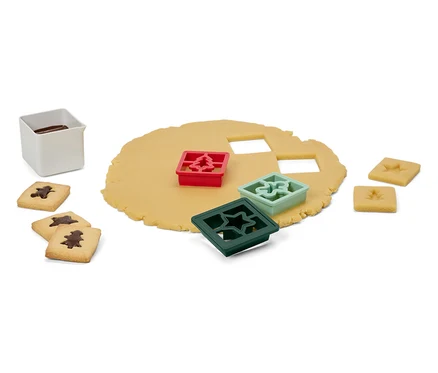 Teig mit Ausstechern in Form von Baum, Stern und Ornament, fertige Kekse und ein weißer Behälter mit Glasur.