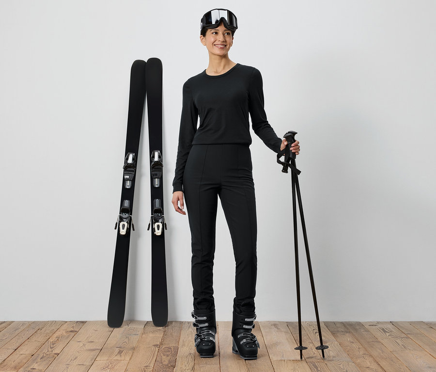 Frau in schwarzer Skibekleidung steht neben Skiern und Stöcken.