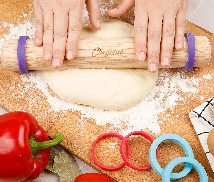 Zwei Hände rollen Teig mit einem Chefclub Nudelholz auf einem Holzbrett aus.
