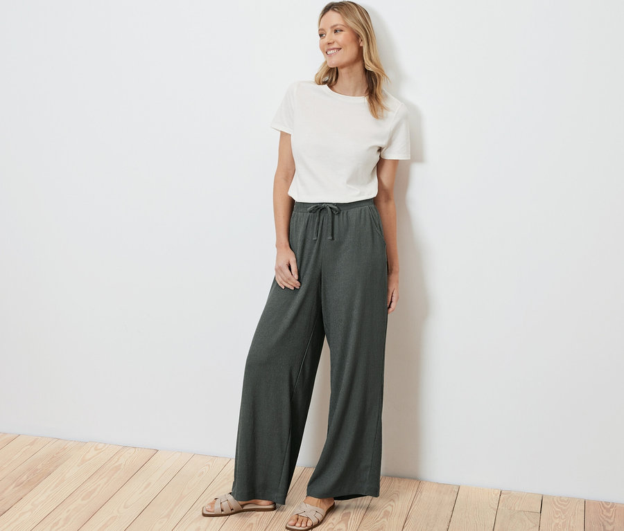 Frau steht und trägt eine grüne Schlupfhose in Crinkle-Optik und ein weißes T-Shirt.