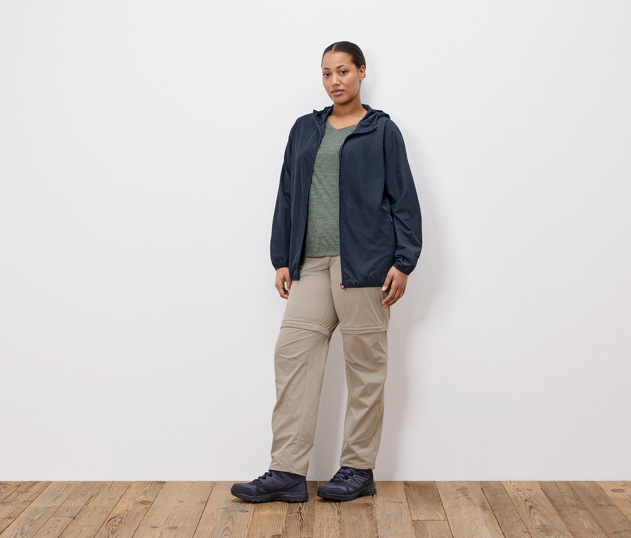 Eine Frau steht vor einer weißen Wand und trägt eine marineblaue Jacke, ein olivgrünes T-Shirt, Khakihosen und marineblaue Schuhe.