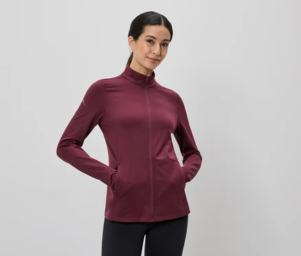 Frau mit Pferdeschwanz trägt eine weinrote Jacke mit durchgehendem Reißverschluss und schwarzen Leggings, die Hände in den Taschen.