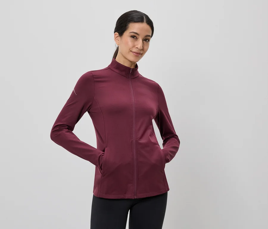 Frau mit Pferdeschwanz trägt eine weinrote Jacke mit durchgehendem Reißverschluss und schwarzen Leggings, die Hände in den Taschen.