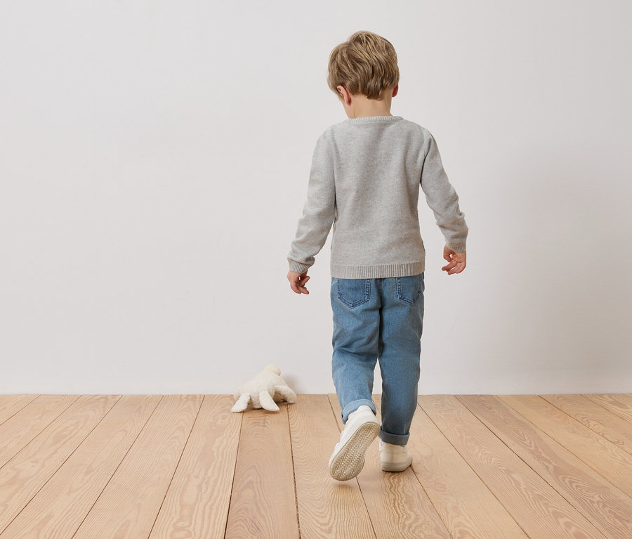 Kind von hinten in grauem Pullover und Jeans, geht auf Holzboden an einer Wand entlang. Ein Stofftier liegt auf dem Boden.