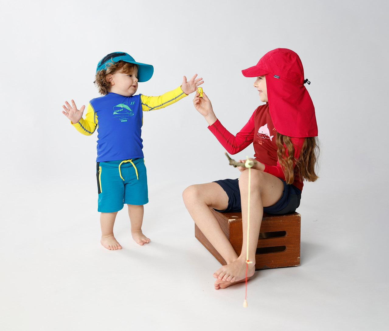 Ein Kind bietet einem sitzenden Kind einen Gegenstand an. Das sitzende Kind trägt eine JAKO-O Kappe mit Nackenschutz, traube.