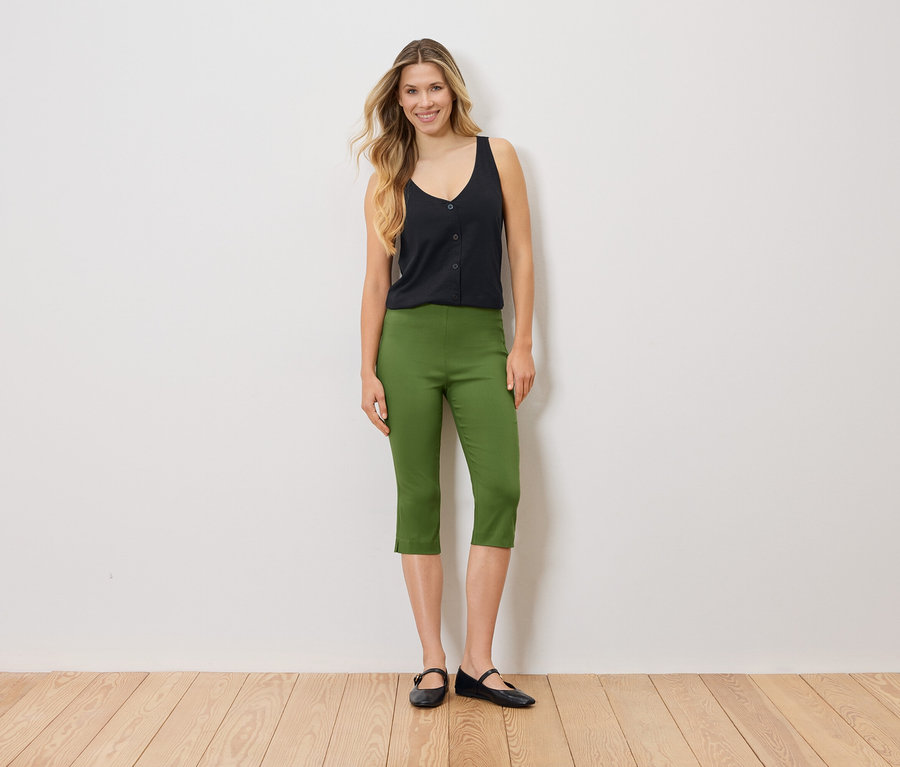 Frau steht vor einer weißen Wand und trägt ein schwarzes Top, eine grüne Caprihose und schwarze Ballerinas.