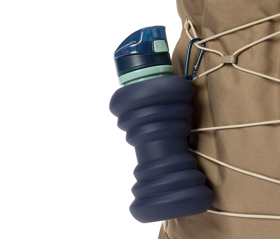 Nahaufnahme einer blauen, zusammendrückbaren Wasserflasche, die mit einem Karabiner an einem beigen Rucksack befestigt ist.